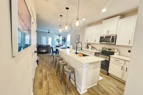 Modern kitchen with white cabinetry, stainless steel appliances, white quartz island with metal bar stools, pendant lights, and open floor plan to living room.
