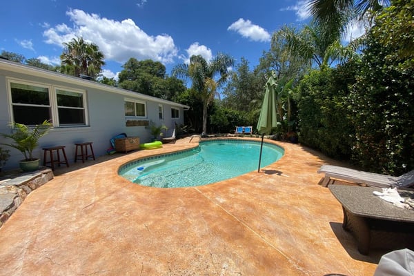 Backyard with kidney-shaped swimming pool, terracotta patio, white house, palm trees, and green umbrella