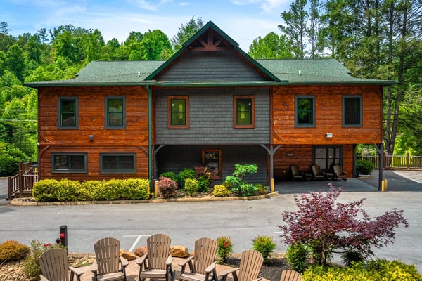 Modern log cabin with green metal roof, covered porch, and Adirondack chairs in landscaped front yard surrounded by trees
