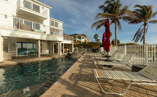 Beachfront house with pool, lounge chairs, red umbrella, palm trees, and white railings under blue sky